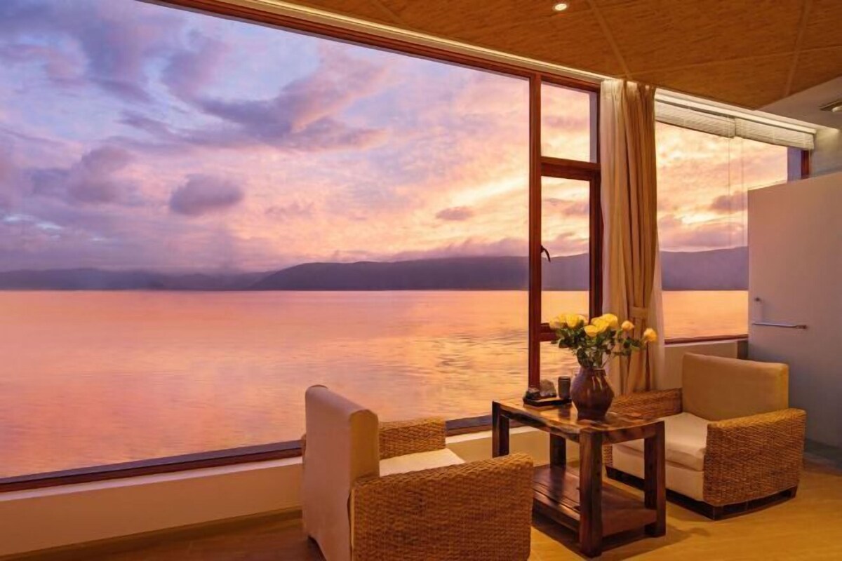 A room with a large window overlooking a body of water, a table with chairs, and a vase with flowers.