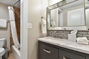 A bathroom with a white toilet, a shower with a white curtain, a sink with a marble countertop, a large mirror, and a towel rack.