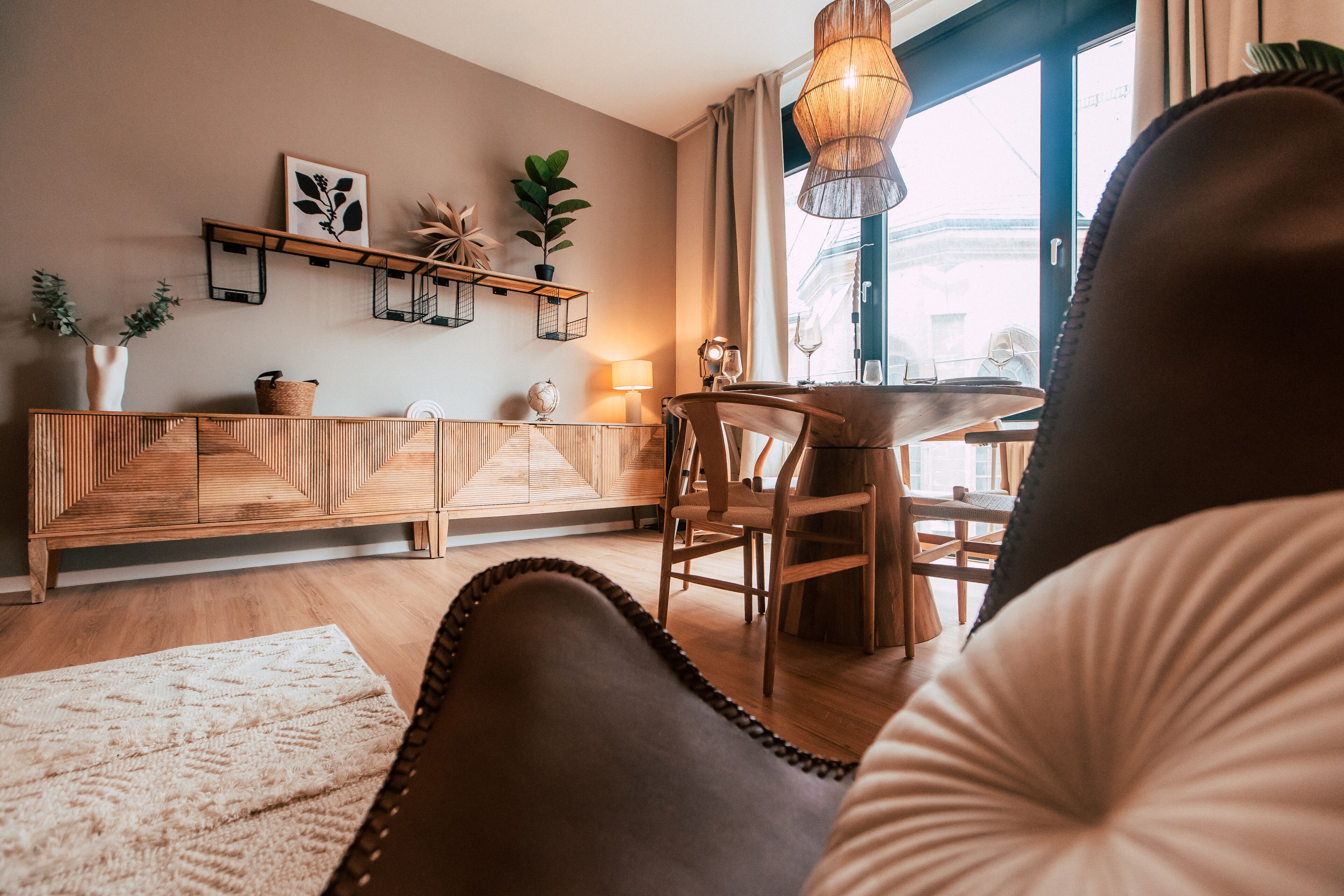 Una sala de estar acogedora con un sofá de madera, una mesa de comedor con sillas, un lámpara colgante y un ventanal con cortinas.