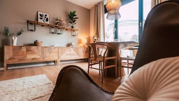 Una sala de estar acogedora con un sofá de madera, una mesa de comedor con sillas, un lámpara colgante y un ventanal con cortinas.