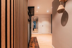 Un pasillo con listones de madera, un banco de madera, un espejo, una puerta y una lámpara de fibra trenzada.