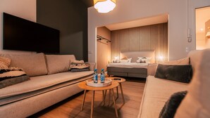 Una camera d'albergo moderna con un divano, un tavolino rotondo con delle bottiglie, un letto con testiera e una TV a parete.