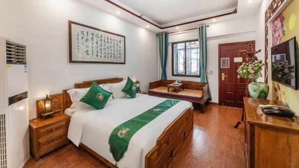 A hotel room with a wooden bed, a bedside table with a lamp, a wall-mounted air conditioner, a framed calligraphy piece, a window with curtains, and a wooden door. - Tongli Defu Caotang Boutique Hostel (Suzhou)