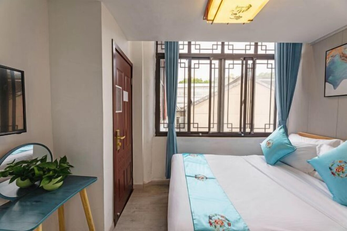 Um quarto com uma cama, uma escrivaninha com uma planta, uma janela com cortinas e uma porta.