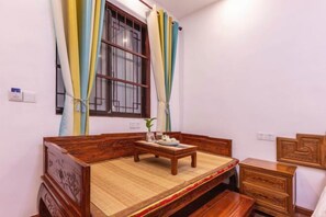 Quarto com cama de madeira, uma mesinha com uma tigela e uma planta, e janela com cortinas.