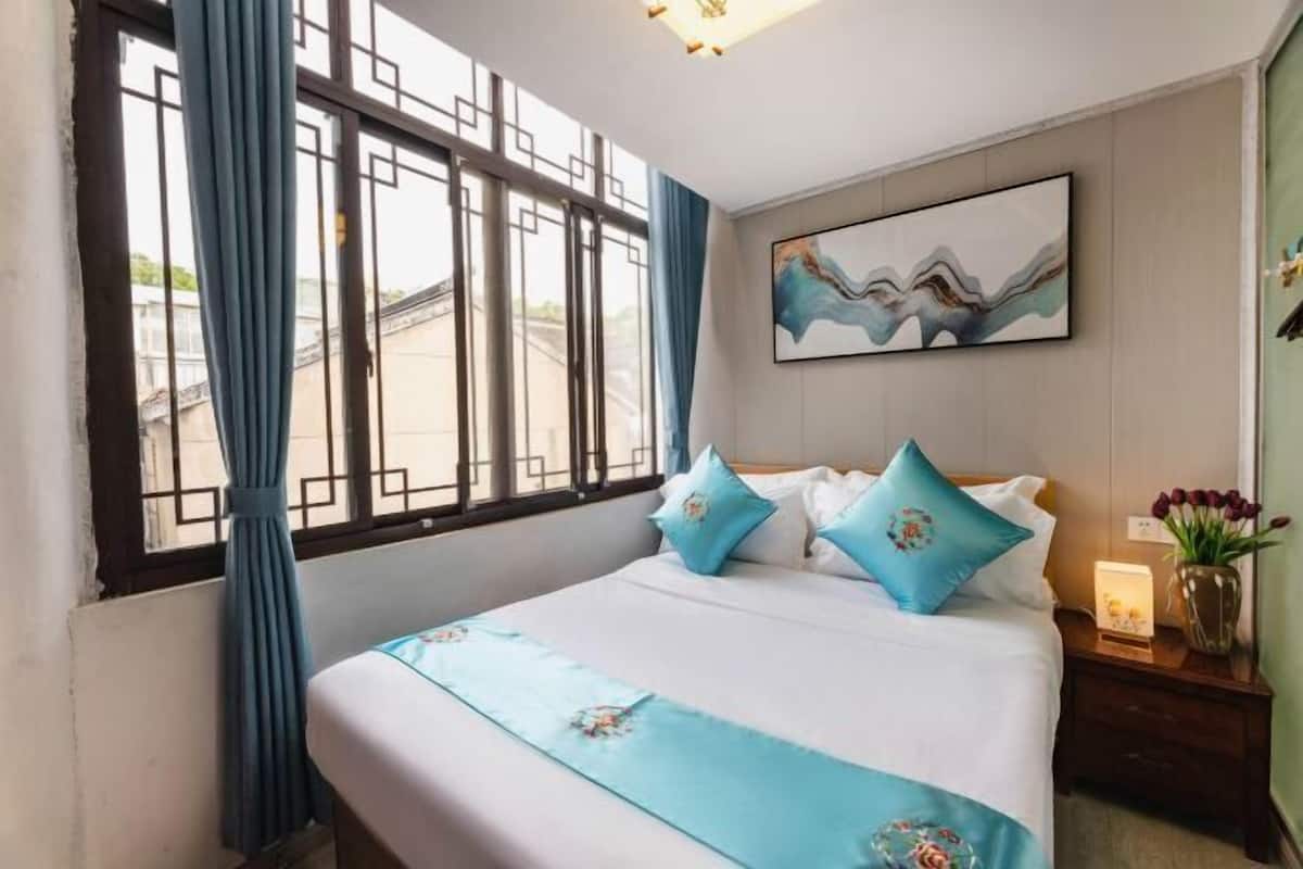 A hotel room with a bed, curtains, a painting, and a nightstand with a vase.