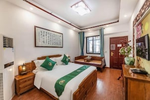 Un dormitorio con cama, mesitas de noche, aire acondicionado instalado en la pared, ventana con cortinas, puerta de madera y una obra de arte enmarcada en la pared.