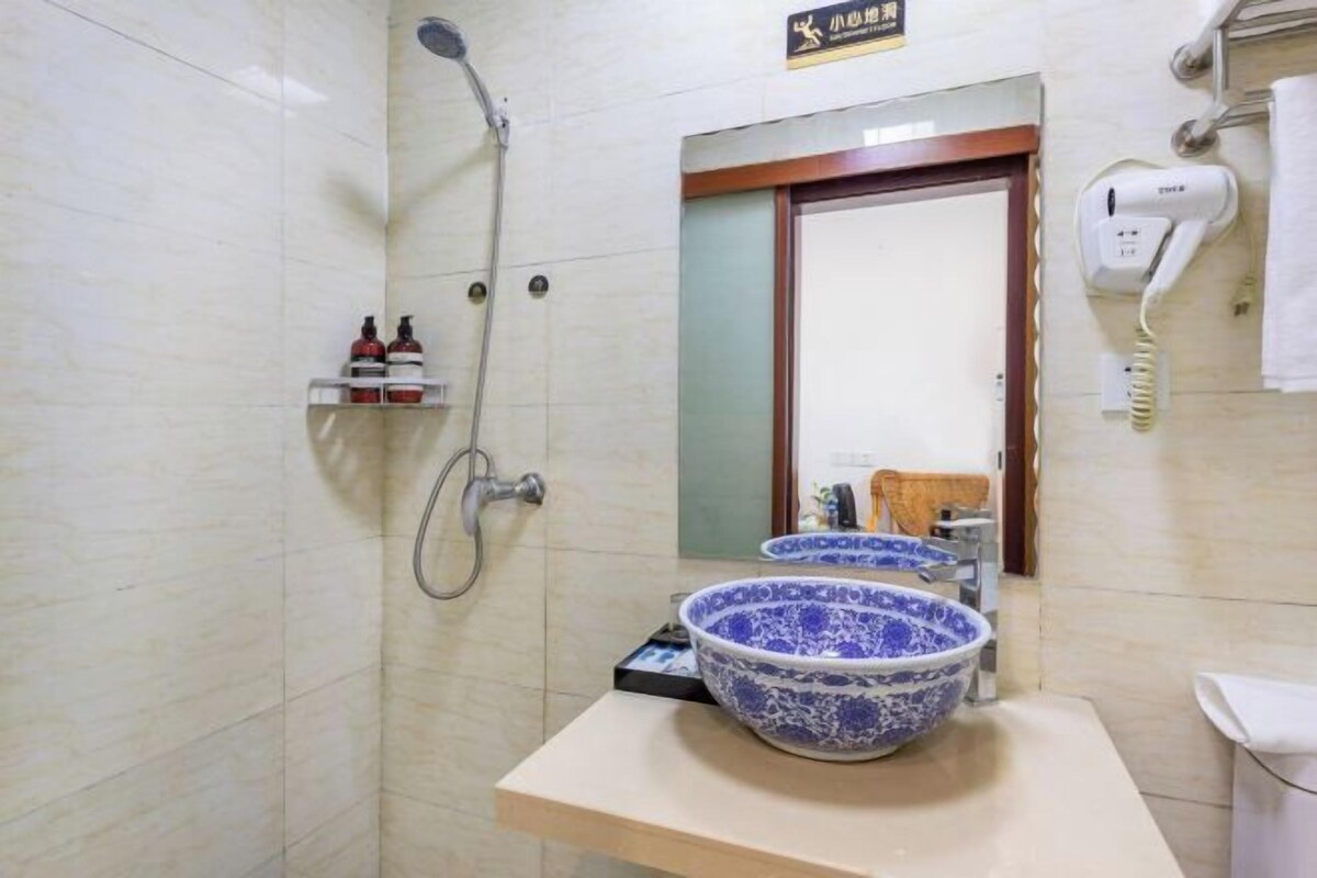 Uma casa de banho com um espelho grande, lavatório azul e branco, chuveiro e secador de cabelo na parede.