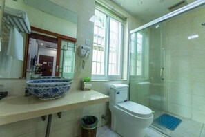 Casa de banho com sanita, cabine de duche envidraçada, lavatório com cuba azul e branca, e janela que permite a entrada de luz natural.