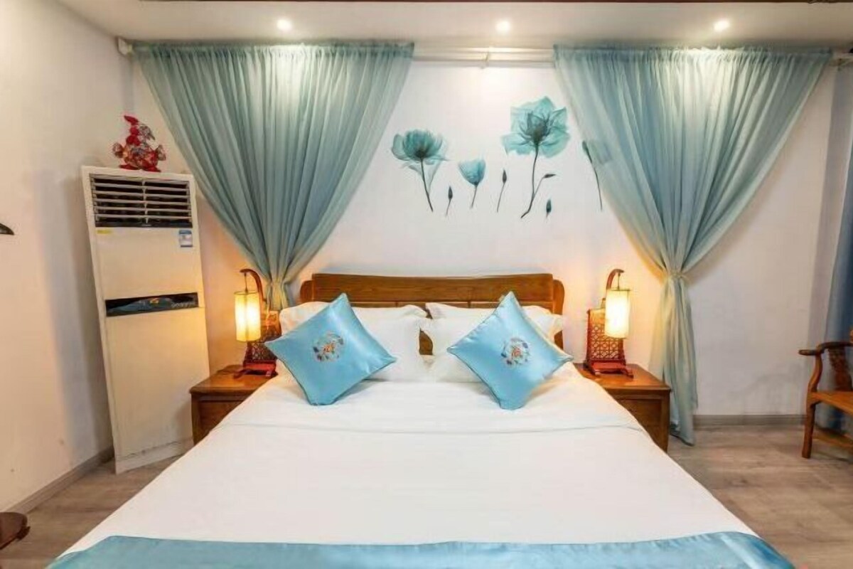 Quarto com cama, duas mesas de cabeceira com abajures, ar-condicionado fixado na parede e um quadro floral.