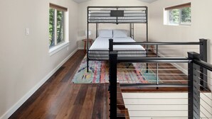 Une chambre avec des lits superposés, un plancher en bois, un tapis coloré et une fenêtre sur le côté gauche.
