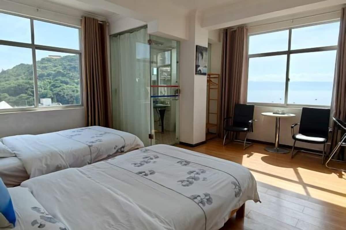 A hotel room with two beds, a small table, two chairs, and large windows with a view of the sea.