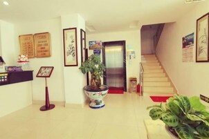 Uma área de recepção de hotel com balcão, obras de arte emolduradas, planta em vaso, escada e porta de acesso ao elevador.