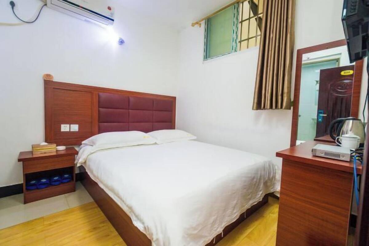 A hotel room with a wooden bed, a bedside table with a lamp, a small shelf with bottles, a mirror, and a window with curtains.