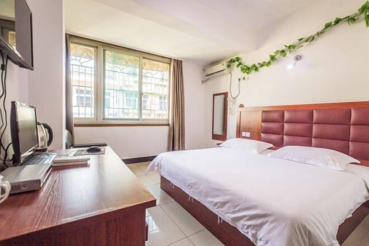 A hotel room with a bed, a desk with a computer, a window, and an air conditioning unit.