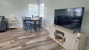 Una sala de estar con televisor, piso de madera, una mesa de comedor con sillas, un sofá y una lámpara.