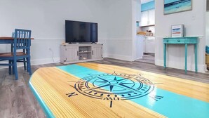 Una habitación con suelo de madera, un diseño de brújula, un televisor, una silla azul, una mesita y una puerta que da a otra habitación.
