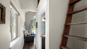 Un couloir étroit avec une échelle en bois, une fenêtre à cadre blanc et une porte donnant sur une salle de bain.