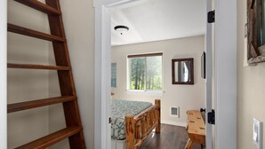 Une chambre avec un lit en bois, une fenêtre donnant sur des arbres, une échelle et une porte menant à une autre pièce.