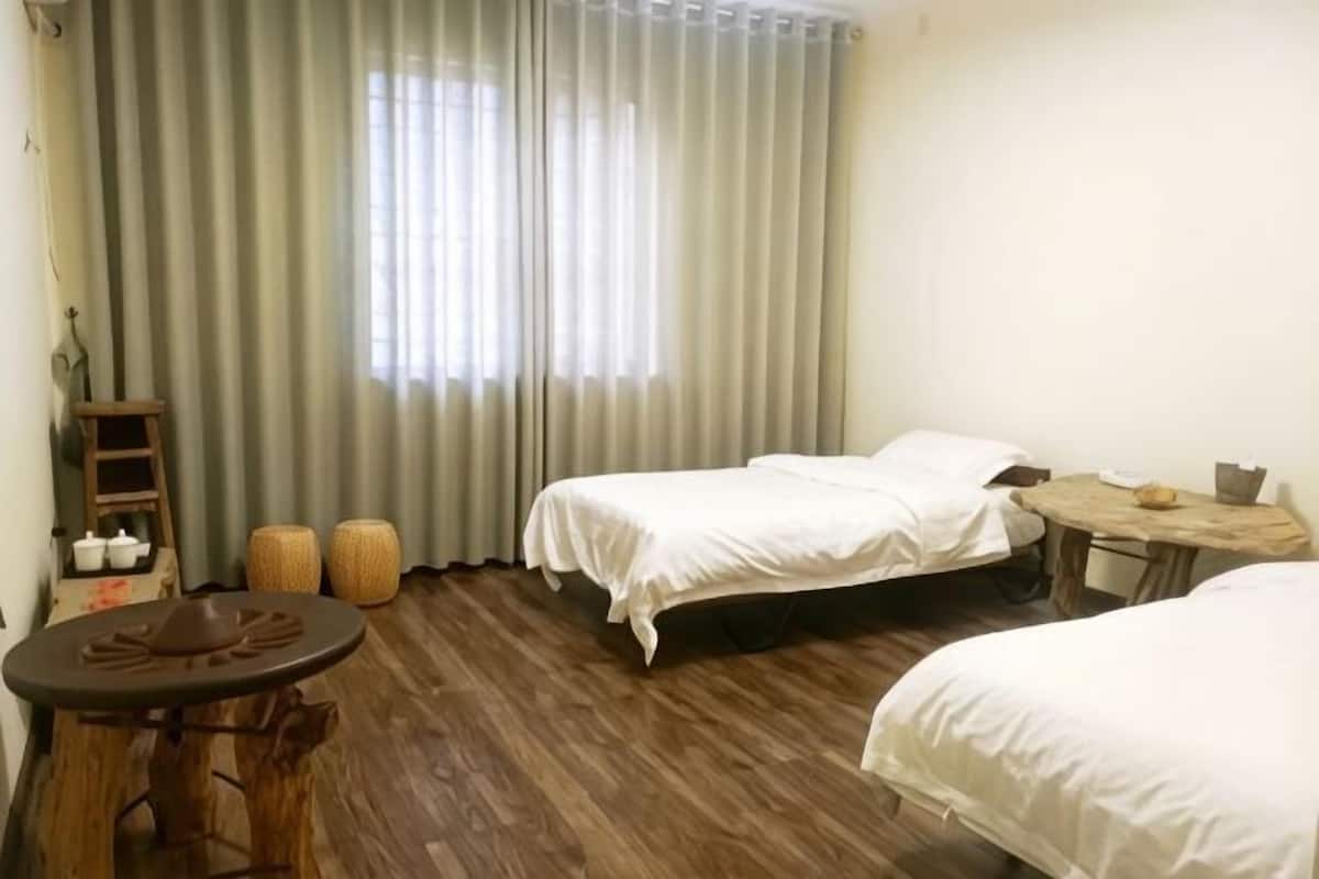 A hotel room with two beds, a small table, a wooden stool, and a window with curtains.