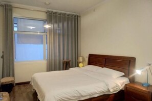 Una habitación de hotel con una cama grande, una mesita de noche con lámpara, una pequeña mesa con lámpara, una ventana con cortinas y piso de madera.