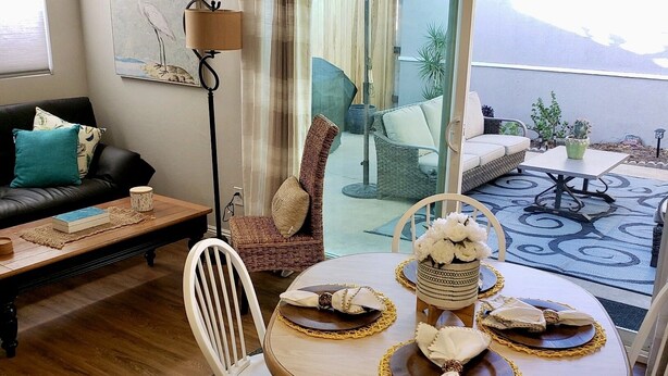 Una sala de estar con sofá, mesa de comedor con sillas, lámpara de pie y vistas al exterior.