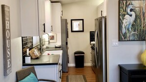 Una cocina moderna con gabinetes blancos, electrodomésticos de acero inoxidable y un pequeño comedor con una silla de mimbre y una mesa.