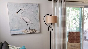 Una sala de estar con un sofá negro, una mesa de centro de madera, un sillón de mimbre, una lámpara de pie y un cuadro de un pájaro.
