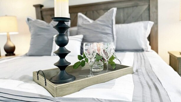 Una cama bien hecha con una bandeja que contiene una vela, vasos y un pequeño jarrón.