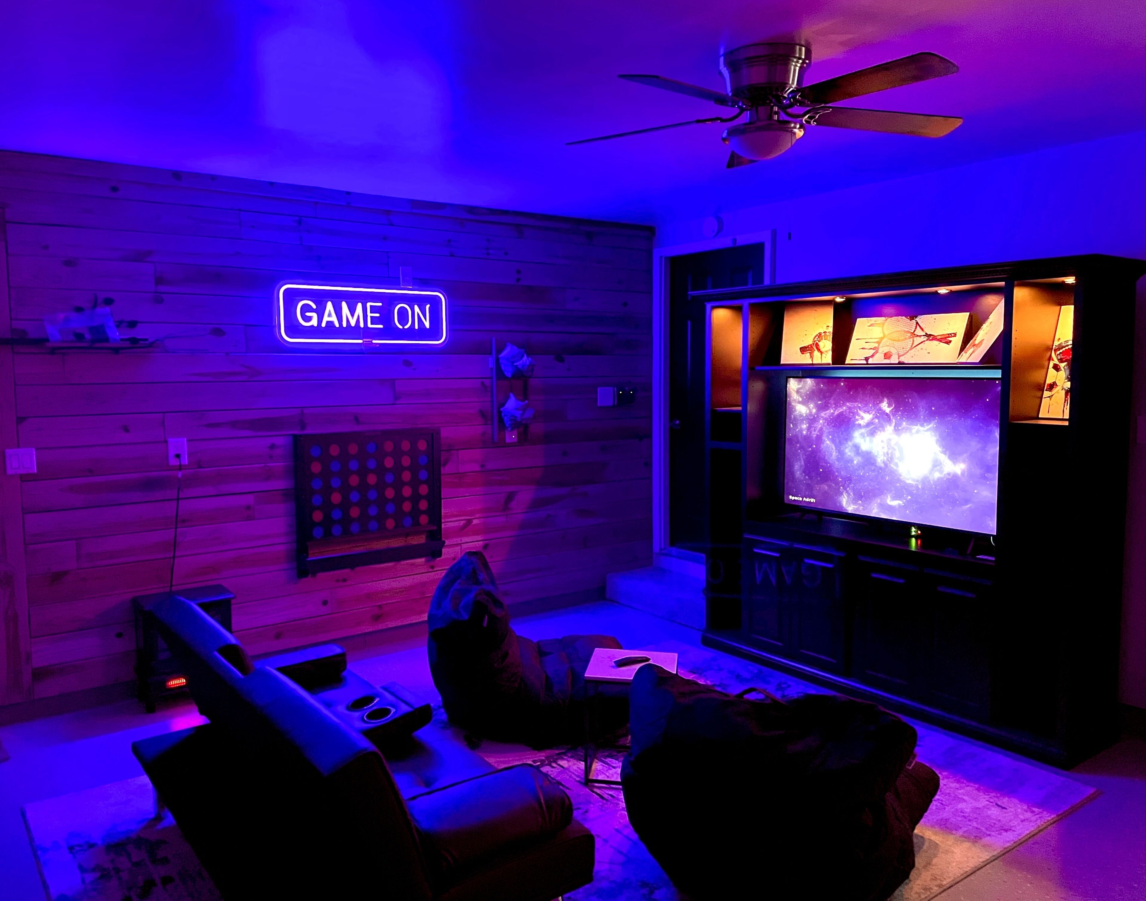 A room with a wooden wall, a ceiling fan, a neon sign that reads 'GAME ON', a large flat-screen TV displaying a space-themed image, and a gaming console.