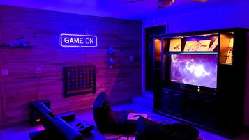 A room with a wooden wall, a ceiling fan, a neon sign that reads 'GAME ON', a large flat-screen TV displaying a space-themed image, and a gaming console.
