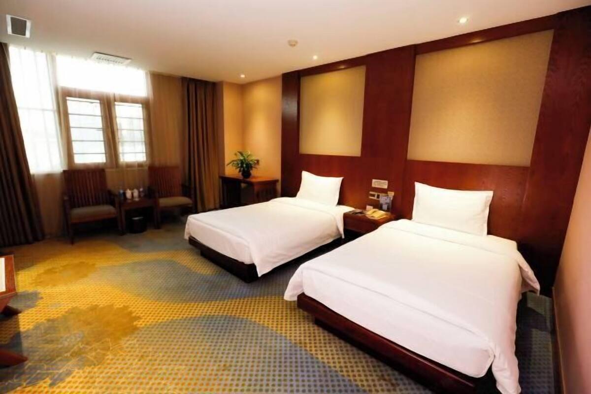 A hotel room with two beds, a desk, a chair, a plant, and a window.