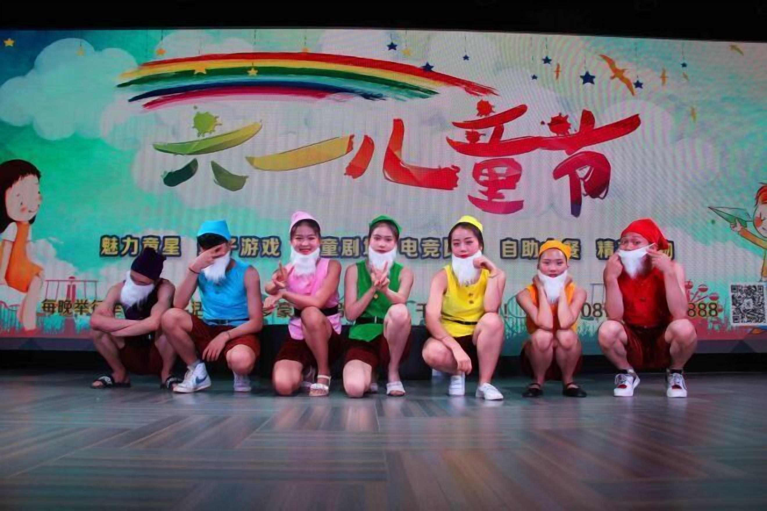 A group of children sitting on stage with a colorful backdrop featuring text and graphics.