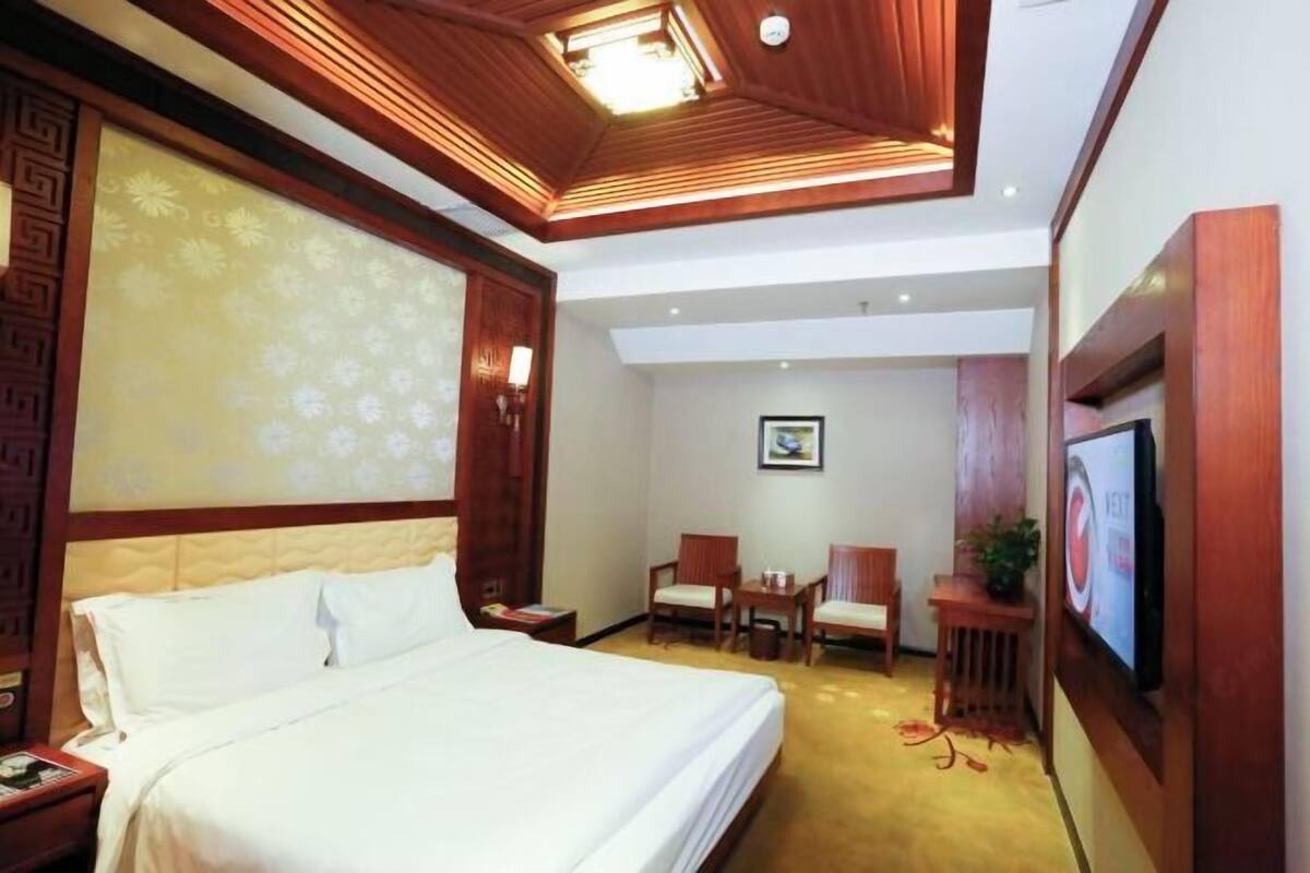 A hotel room with a large bed, a desk with two chairs, a television, and a decorative ceiling.