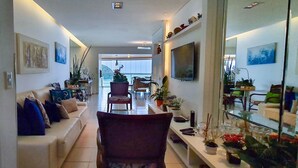 A modern living room with a sofa, chairs, a coffee table, and a TV. There are plants and decorative items on shelves.