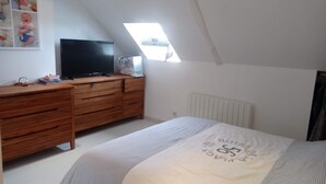 Ein Schlafzimmer mit schrägverdeckter Decke, einer Holzkommode, einem Fernseher und einem Bett mit weißer Bettdecke.