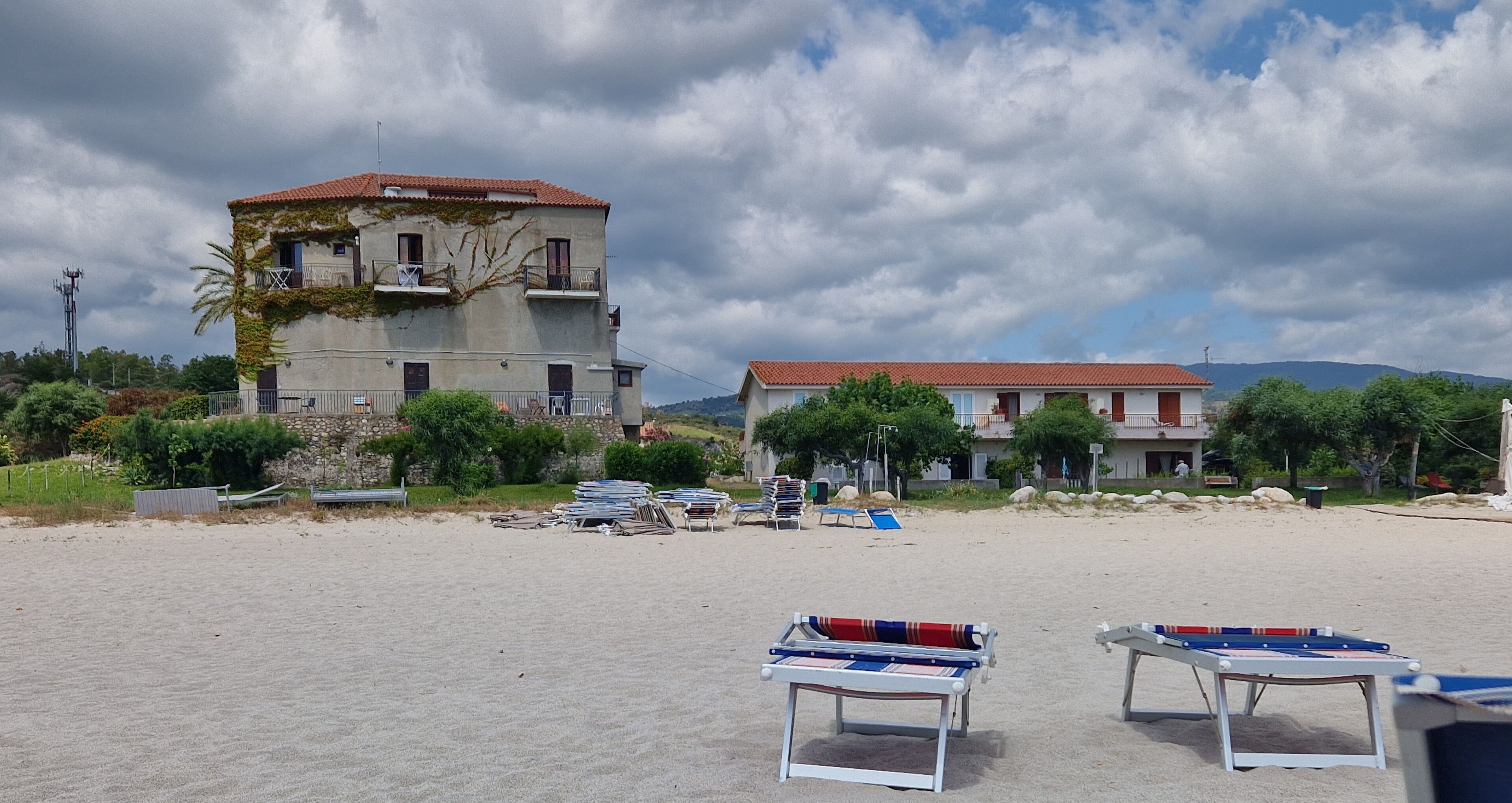 A beach with two buildings, one with a red roof and another with a white roof. There are lounge chairs and a table with a table tennis table in the foreground.