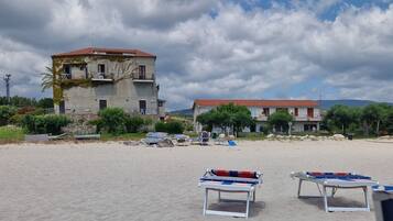 A beach with two buildings, one with a red roof and another with a white roof. There are lounge chairs and a table with a table tennis table in the foreground.