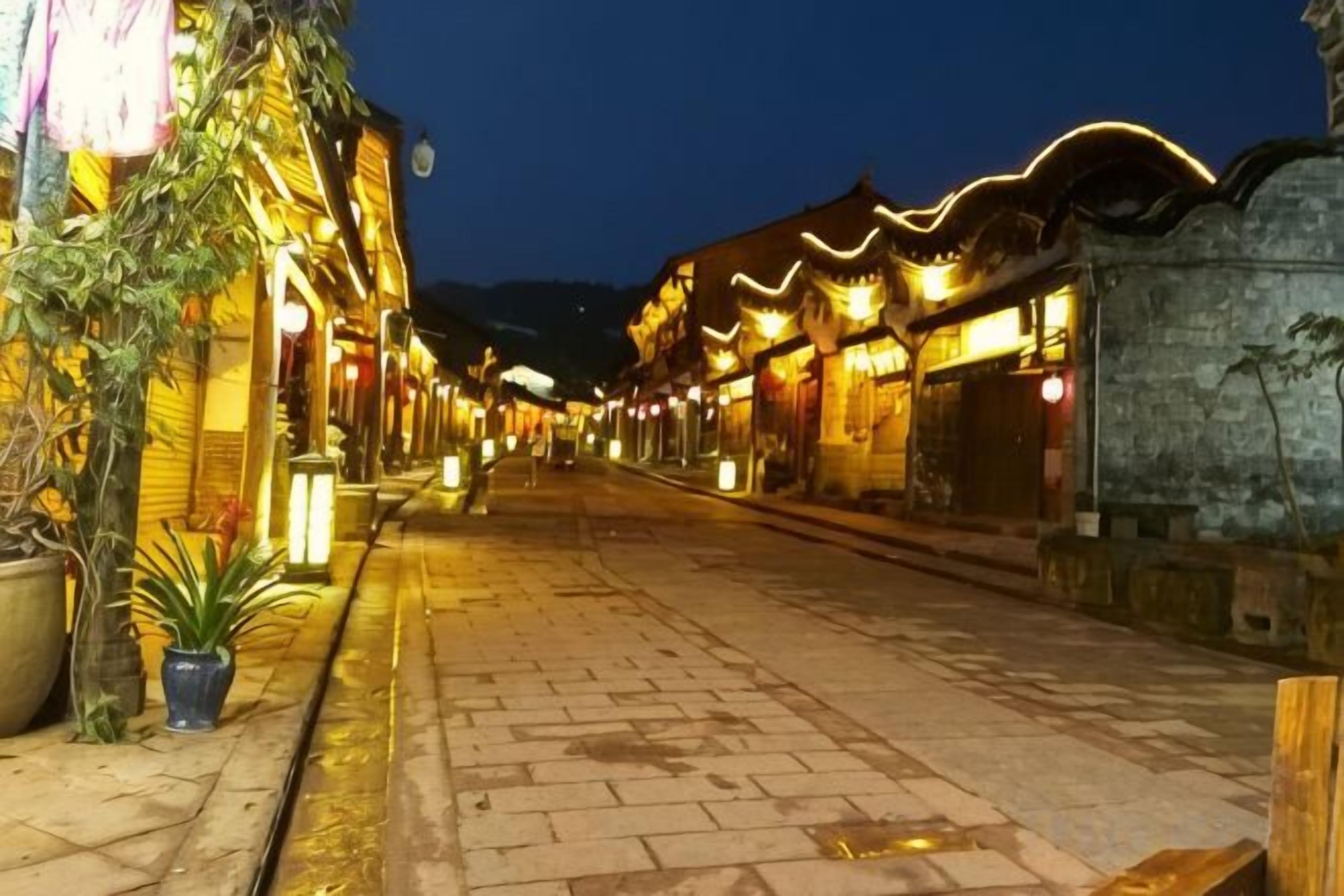 A paved street lined with traditional buildings, some with illuminated signs, and a clear sky.