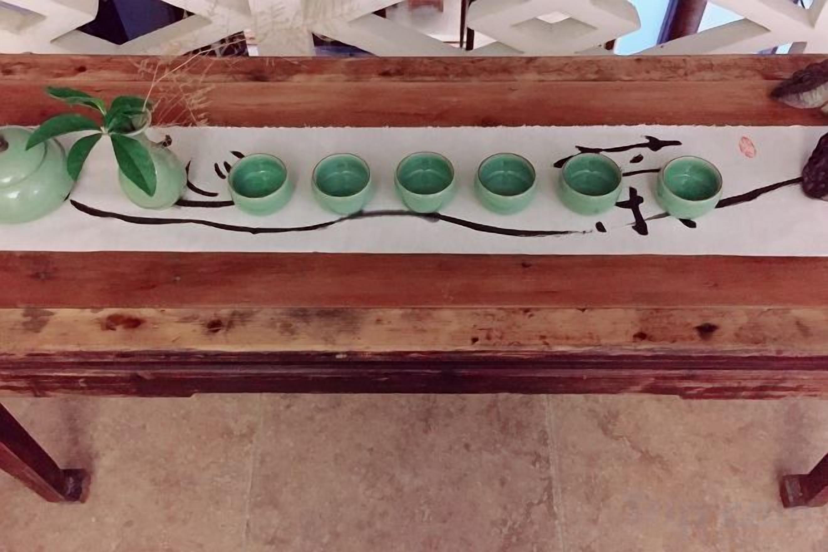 A wooden table with a green vase and six green cups aligned on a white surface.