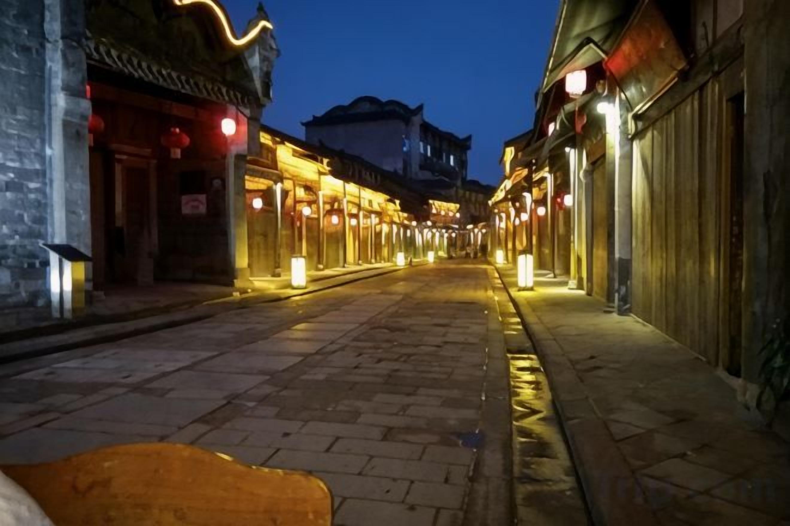 A narrow street with traditional architecture, illuminated by warm lighting, and a clear sky.