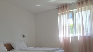 A bedroom with a wooden bed, white bedding, a window with sheer curtains, and a light switch on the wall.