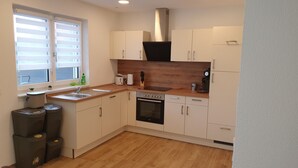 A compact kitchen with a sink, stove, oven, and dishwasher. There is a window with blinds, a countertop with a knife block, and a trash bin.