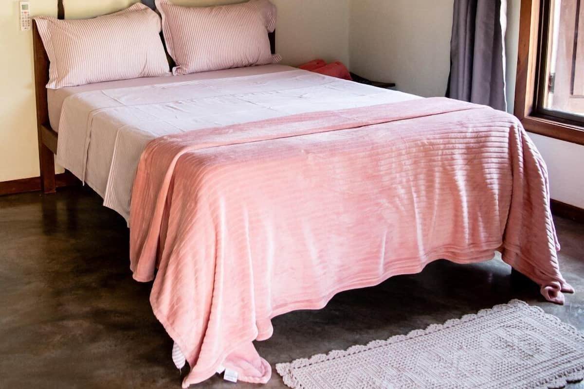 Un dormitorio con cama, almohadas, una mesita de noche, un interruptor de luz en la pared y una ventana con cortinas.