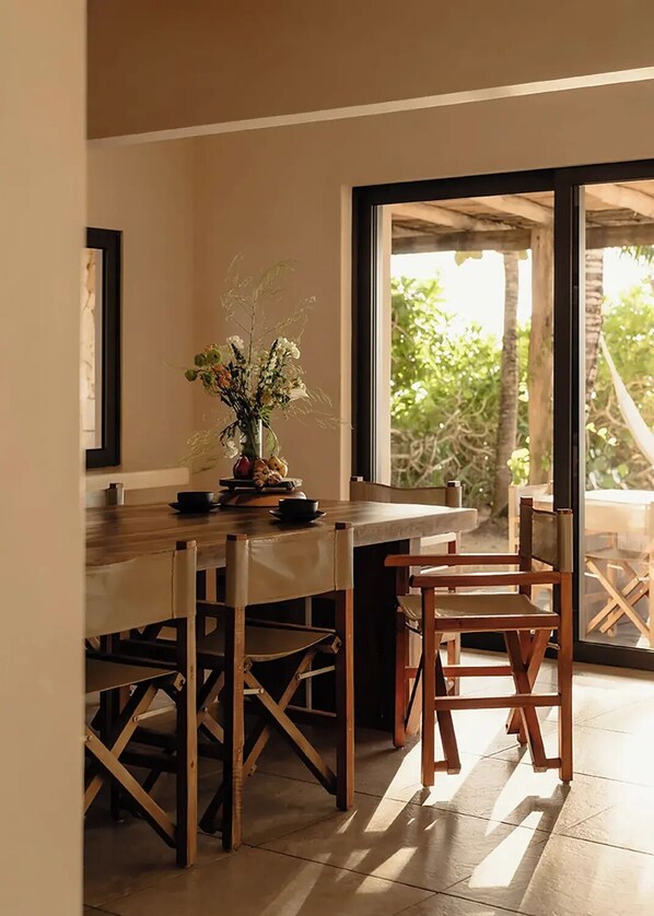 Un comedor con una mesa y sillas de madera, un jarrón con flores y vista a zonas verdes a través de un amplio ventanal.