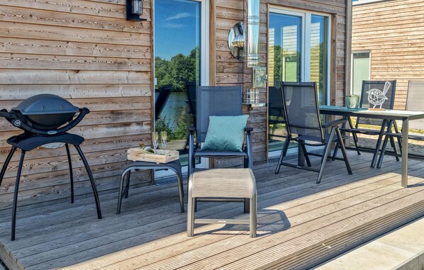 Eine Terrasse im Freien mit einem Grill, zwei Stühlen, einem kleinen Tisch mit Flasche und Gläsern sowie einer dekorativen Vogelstatue.