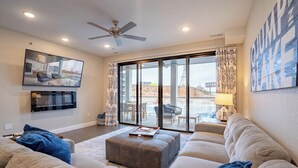 A modern living room with a large flat-screen TV, comfortable seating, and a view of the outdoors through large windows.