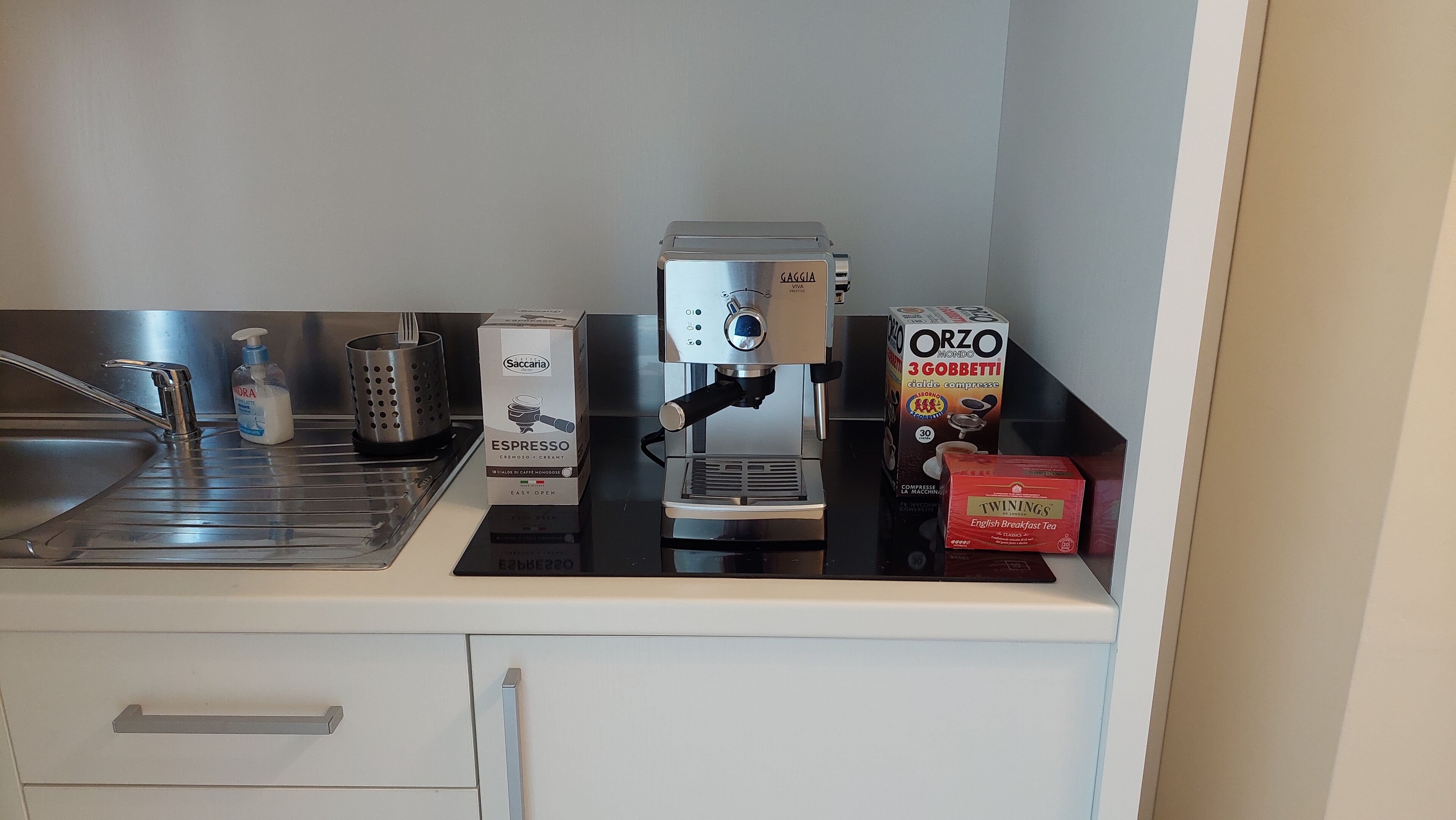 A kitchen counter with a coffee machine, a box of Saccaria espresso, a box of Orzo Mondo, a box of Twinings English Breakfast Tea, a bottle of hand soap, and a metal grater.