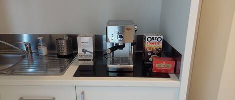 A kitchen counter with a coffee machine, a box of Saccaria espresso, a box of Orzo Mondo, a box of Twinings English Breakfast Tea, a bottle of hand soap, and a metal grater.