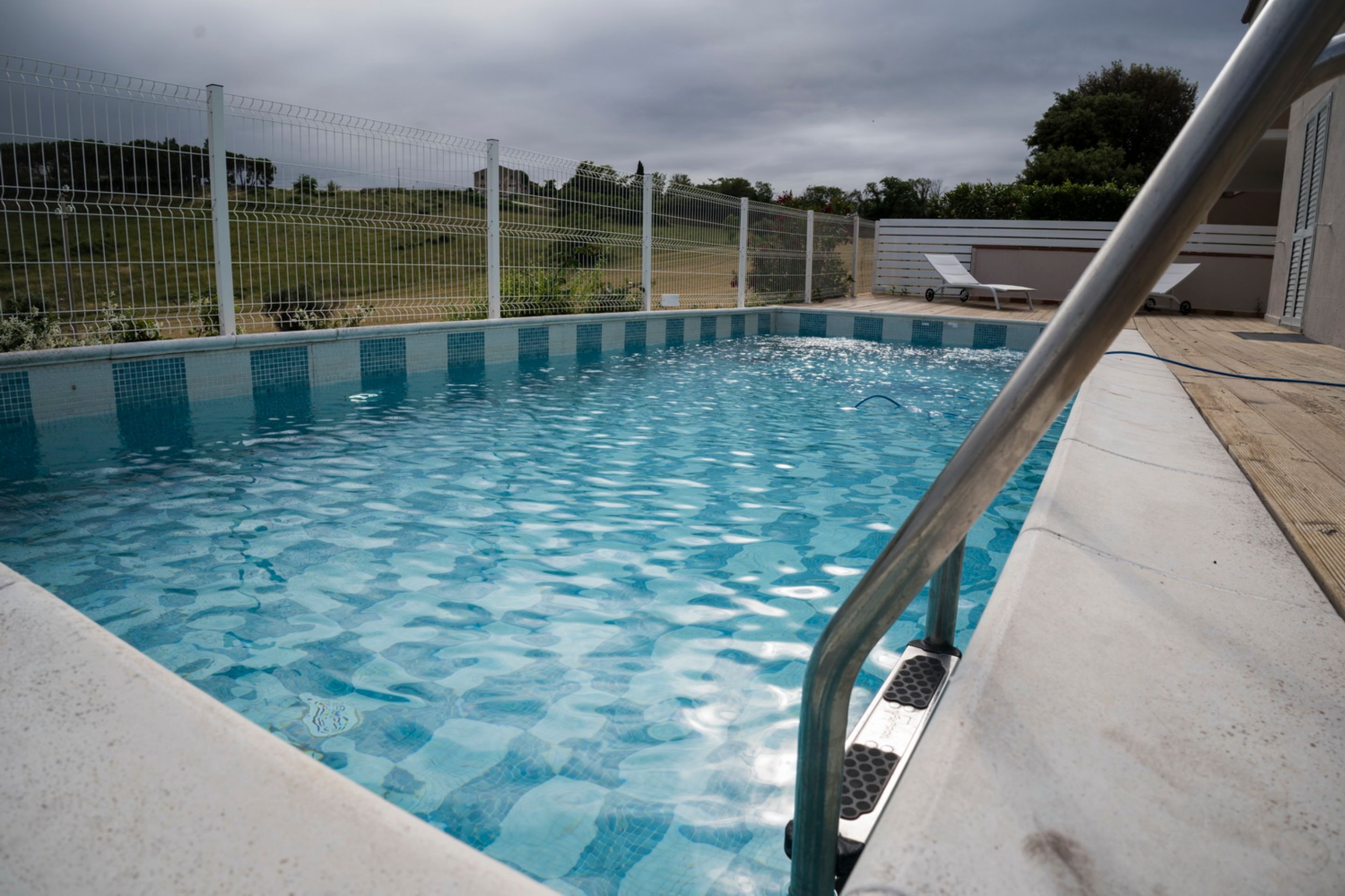 Una piscina con barandilla metálica de seguridad, rodeada por una valla y un área de descanso con sillas.